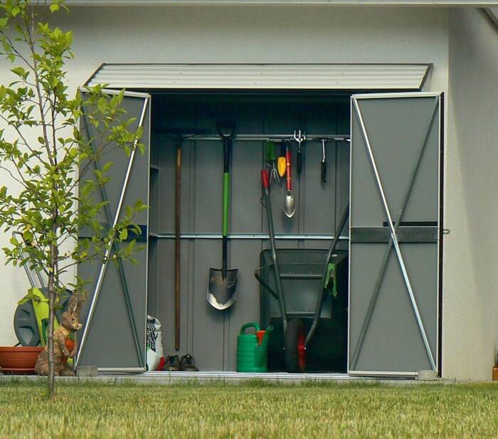 Prosty metalowy domek narzędziowy postawiony przy ścianie budynku mieszkalnego. Dzięki dwuskrzyd ...