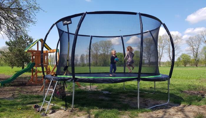 Duża trampolina ogrodowa z bawiącymi się dziećmi na tle zielonego pola. Wewnętrzna siatka oraz o ...